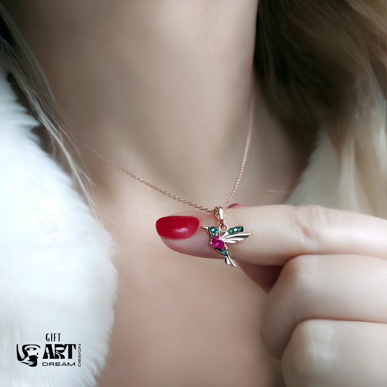 May include: A rose gold necklace with a hummingbird pendant. The pendant features a hummingbird design with red and green gemstones. The necklace is displayed against a white background.