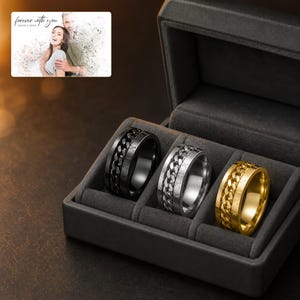 May include: Three rings in black, silver, and gold tones, each featuring a central rotating chain link band and Roman numeral engravings, displayed in a dark grey velvet-lined box. A small card shows a couple with the text forever with you.