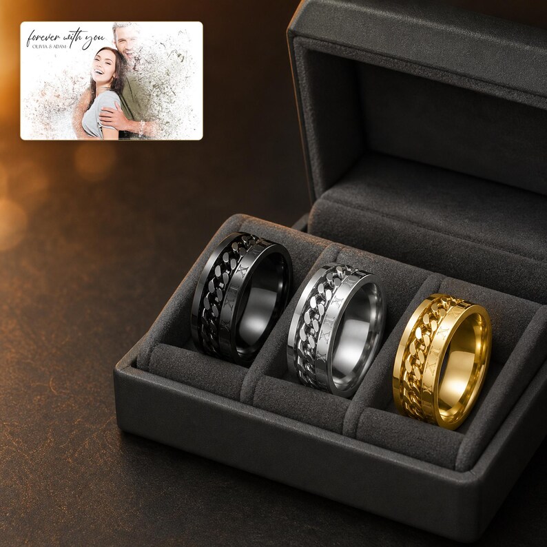 May include: Three rings in black, silver, and gold tones, each featuring a central rotating chain link band and Roman numeral engravings, displayed in a dark grey velvet-lined box. A small card shows a couple with the text forever with you.