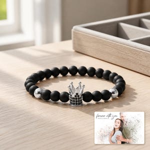 Puede incluir: Una pulsera de cuentas con cuentas esf&eacute;ricas de color negro mate, dos cuentas esf&eacute;ricas de tono plateado pulido y un dije central de corona de tono plateado incrustado con peque&ntilde;as piedras negras. Una peque&ntilde;a tarjeta rectangular muestra el texto "forever with you".
