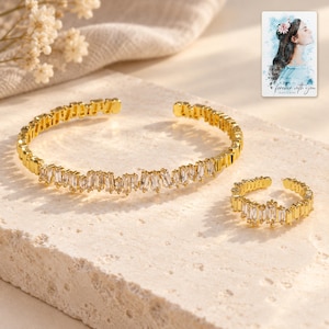 Puede incluir: Un brazalete abierto de tono dorado y un anillo abierto a juego, ambos con una serie de piedras transparentes de corte baguette dispuestas en un patrón irregular y dentado. Se incluye una pequeña tarjeta rectangular con una ilustración en acuarela y el texto "forever with you".