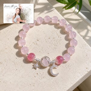 Puede incluir: Una pulsera de cuentas con esferas de cuarzo rosa claro, dos cuentas craqueladas de color rosa oscuro y espaciadores plateados. La pulsera incluye una cuenta central de cuarzo rosa en una jaula plateada, un dije de estrella y uno de luna creciente. Una tarjeta dice forever with you.
