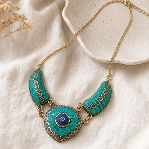 May include: A gold-toned metal necklace featuring a central hexagonal pendant and two curved side pieces, all inlaid with small turquoise-colored mosaic tiles and accented with circular dark blue stone insets throughout the design.