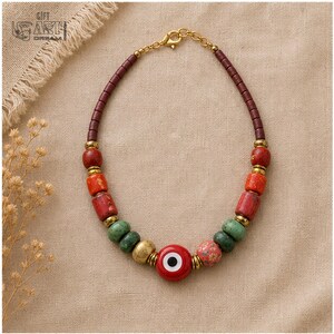 Puede incluir: Un collar con una cuenta central roja de mal de ojo, flanqueada por cuentas verdes, naranjas y con motivos florales, con cuentas cil&iacute;ndricas de color marr&oacute;n oscuro que se extienden hasta un cierre de metal en tono dorado.