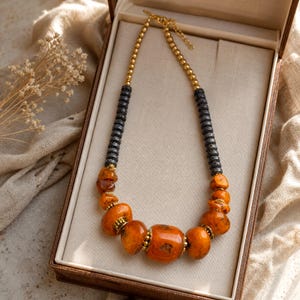 May include: A necklace featuring a strand of polished gold-tone beads, followed by segments of dark grey textured cylindrical beads, and a central arrangement of large, smooth, irregular orange amber-coloured stones accented with small gold-tone spacers.