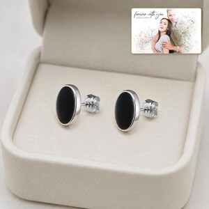 Black oval stud earrings with silver-tone edges, presentation in an open jewelry box. A personalized greeting-card example appears in the corner.