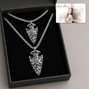 May include: Two silver-toned arrowhead pendants featuring engraved Vegvisir symbols, each suspended on a matching braided chain. The necklaces are presented inside a black gift box alongside a small rectangular card displaying a photograph of a couple.