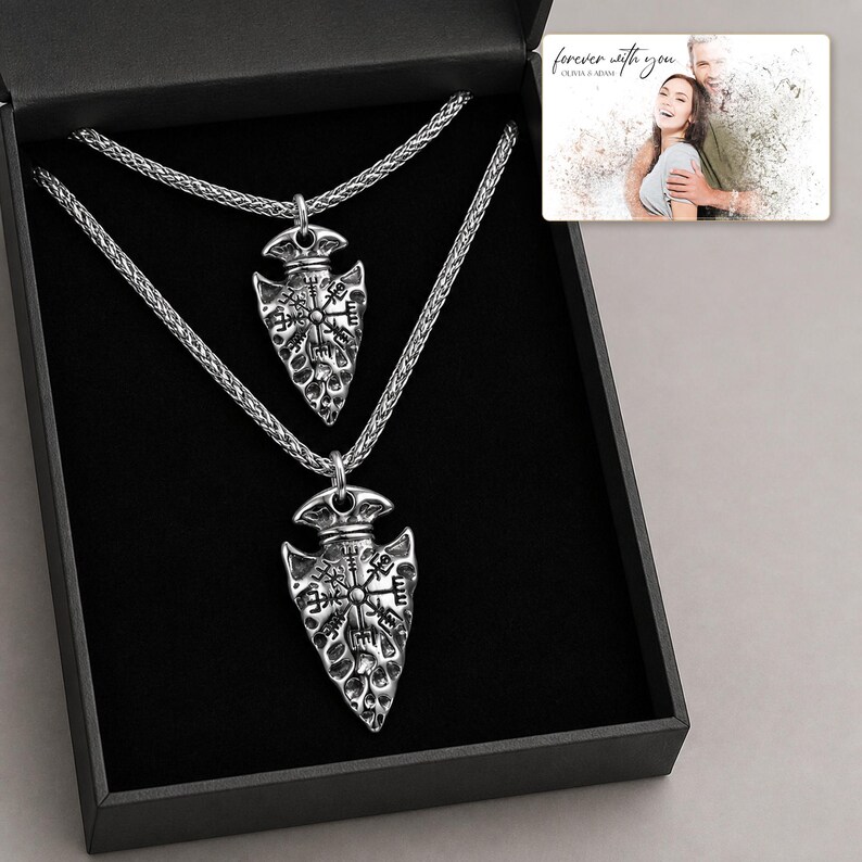 May include: Two silver-toned arrowhead pendants featuring engraved Vegvisir symbols, each suspended on a matching braided chain. The necklaces are presented inside a black gift box alongside a small rectangular card displaying a photograph of a couple.