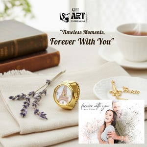 May include: A gold ring watch with a white face showing the Eiffel Tower. The watch sits on a white cloth with lavender and a small plate with a gold Eiffel Tower charm and the word "Paris". A cup of tea and a book are also visible in the image.