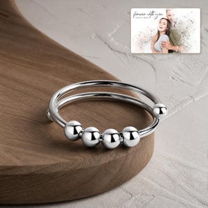 May include: A silver-toned metal ring featuring a double band design with four smooth, round beads that slide along the wire. In the upper right corner, a rectangular card displays a photograph of a couple with the text "forever with you".