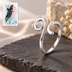 Polished silver-tone cat ring displayed on charcoal slate with the pink ears visible. A personalized digital greeting card is shown alongside.