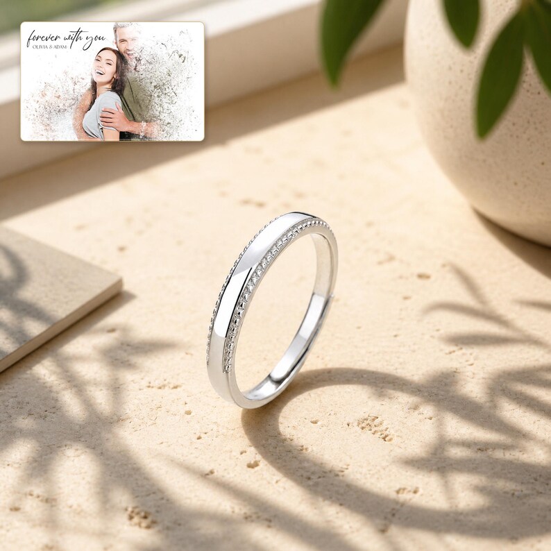 May include: A silver-toned ring featuring a smooth, polished center band flanked by two rows of small, clear stones. In the upper corner, a rectangular print displays a couple with the text "forever with you" and "OLIVIA & ADAM".