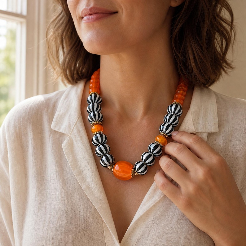 Single-strand orange and black-and-white striped bead statement necklace with gold-tone spacers and a large orange oval center bead displayed on a jewelry bust with the full bead sequence visible