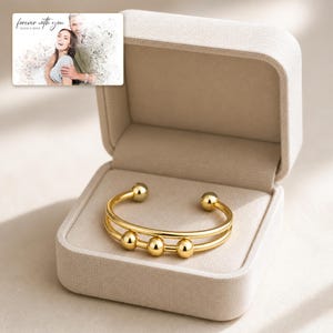 May include: A gold-toned double-band cuff bracelet featuring three small sliding spheres along the front, presented in an open beige fabric-covered jewelry box. A small rectangular card with a photo of a couple and the text forever with you is included.