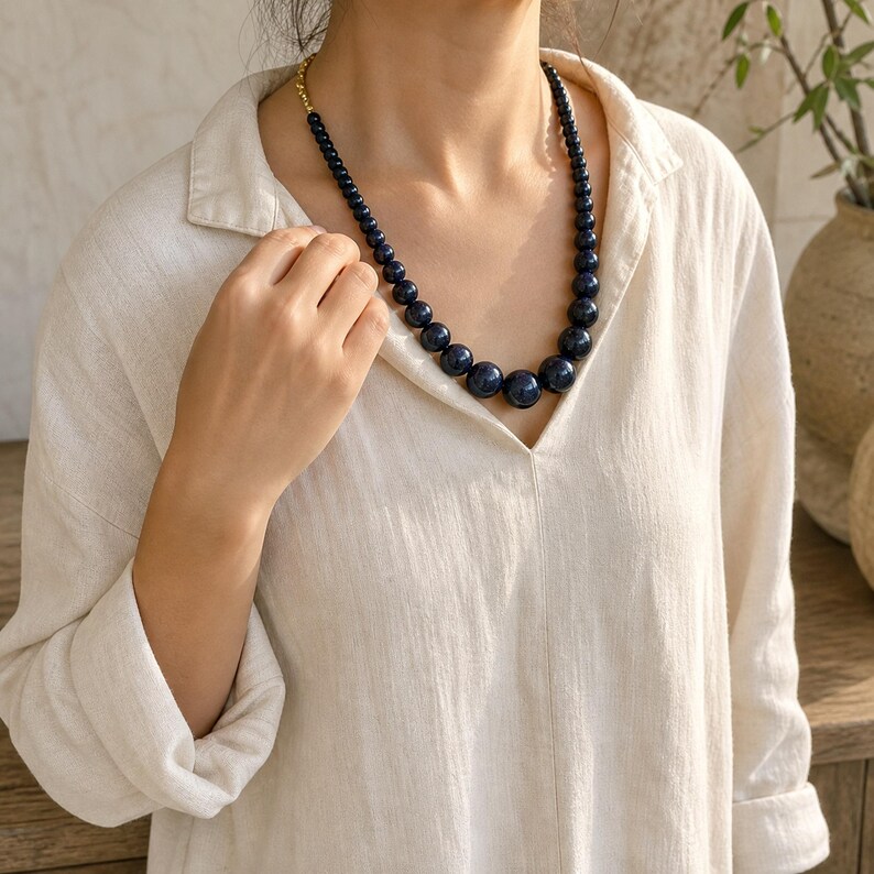 May include: A necklace featuring a strand of graduated dark blue spherical beads. The beads increase in size towards the centre of the strand. The necklace is secured with a gold-toned metal clasp visible at the side of the neck.