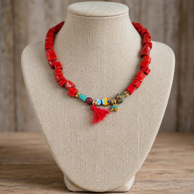 Op de afbeelding: Een ketting met een reeks rode, kubusvormige kralen, geaccentueerd met een kleine blauw-gele visbedel, een rode stoffen kwastje, een kraal met patroon en kleine turquoise kralen, allemaal verbonden door goudkleurige metalen tussenstukjes.
