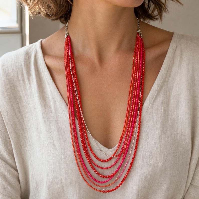 May include: A multi-strand necklace featuring numerous layers of small, round beads in varying shades of red, orange, and pink. The strands are connected at the top by silver-toned metal components, creating a cascading effect that rests against the chest.