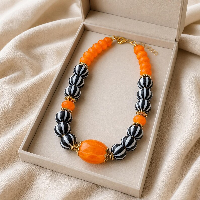 May include: A beaded necklace featuring alternating orange and black-and-white striped spherical beads. The design includes a large, central oval orange bead, gold-toned metal spacers between the beads, and a gold-toned lobster claw clasp closure.
