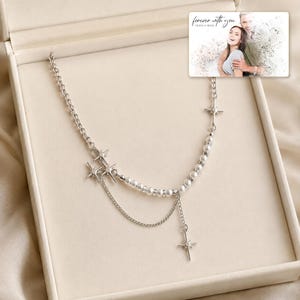 Silver-tone layered necklace with pale beads and pointed star charms, presentation in an open jewelry box. A personalized greeting-card example appears in the corner.
