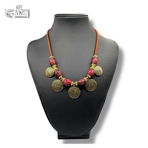May include: A necklace featuring a brown cord, red porous beads, and gold-toned metal spacers and circular pendants with a hammered texture. The necklace is displayed on a black bust. The text GIFT ART DREAM is visible in the upper left corner.