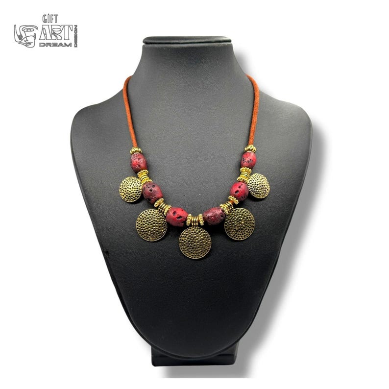 May include: A necklace featuring a brown cord, red porous beads, and gold-toned metal spacers and circular pendants with a hammered texture. The necklace is displayed on a black bust. The text GIFT ART DREAM is visible in the upper left corner.