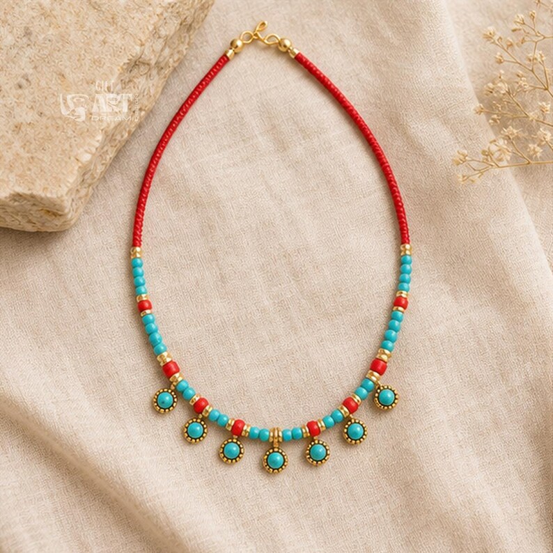 May include: A beaded necklace featuring a vibrant pattern of small red and turquoise beads accented with gold-toned spacers. The lower section includes five dangling circular turquoise charms framed in gold, finished with a gold-toned clasp.