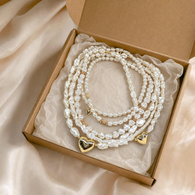 Puede incluir: Un collar de varias hebras con perlas blancas irregulares y dijes dorados en forma de coraz&oacute;n. El collar est&aacute; enrollado dentro de una caja de cart&oacute;n cuadrada forrada con papel de seda blanco arrugado, mostrando un dise&ntilde;o en capas con detalles met&aacute;licos.