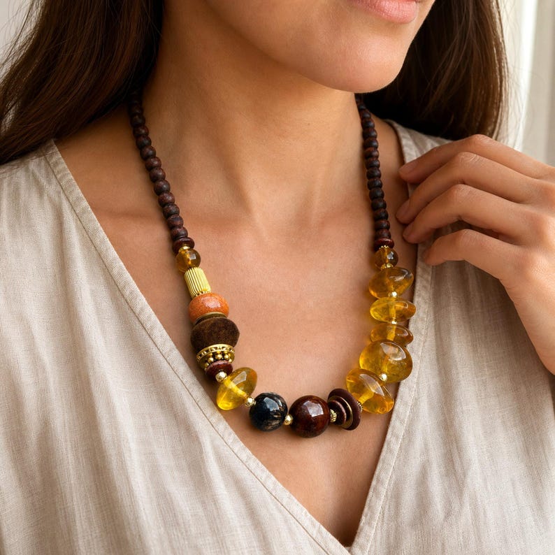Puede incluir: Un collar que presenta una mezcla de cuentas de madera marr&oacute;n oscuro, piedras de resina transl&uacute;cida color &aacute;mbar, una cuenta naranja texturizada, un espaciador cil&iacute;ndrico dorado y una cuenta jaspeada azul oscuro, ensartadas en un cord&oacute;n.