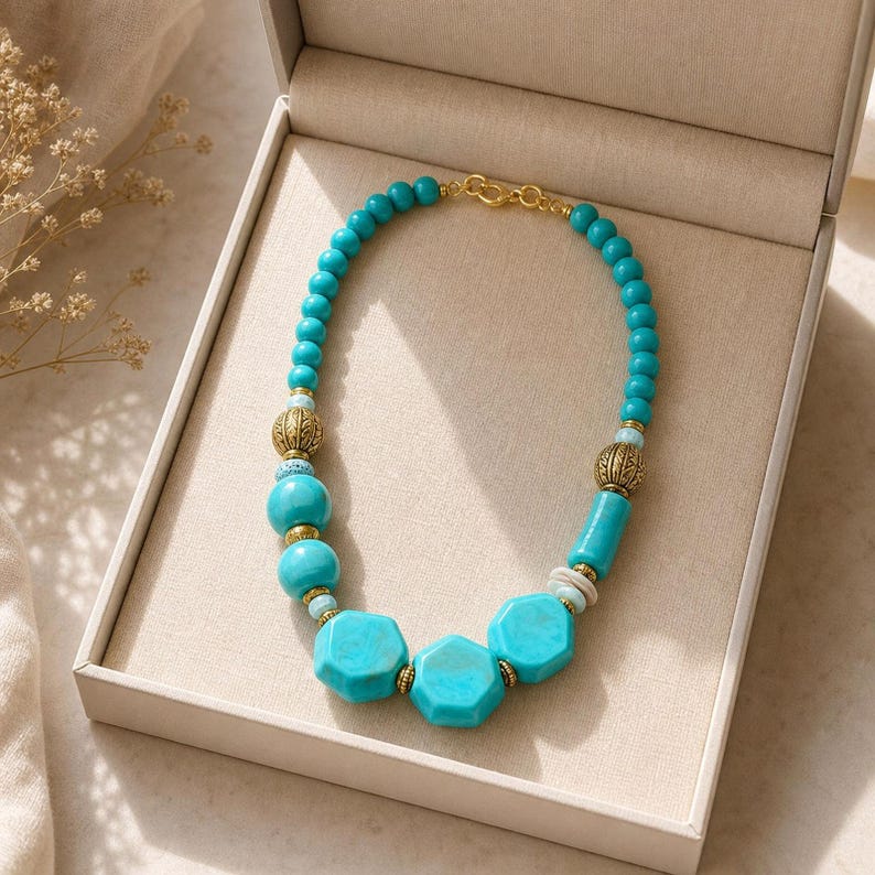 May include: A necklace featuring turquoise-coloured beads in various shapes, including round, cylindrical, and hexagonal, interspersed with ornate gold-toned metal spacers. The piece is finished with a gold-toned metal clasp and is presented inside a beige box.