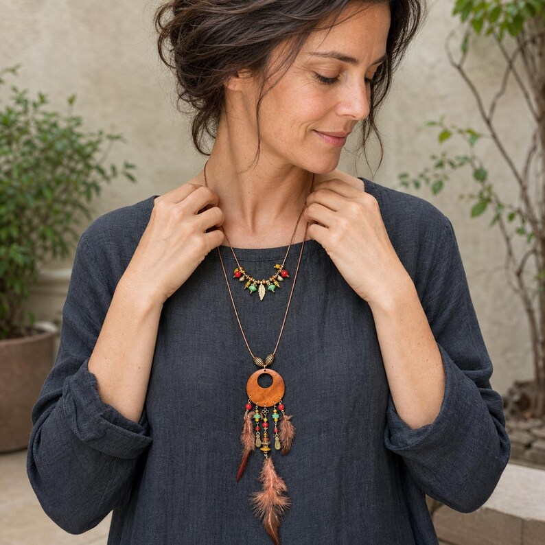 May include: A layered necklace featuring a short strand with small red, green, and gold beads and a long strand with a large circular wooden pendant, dangling metal charms, colorful beads, and brown feathers, worn over a dark gray linen shirt.