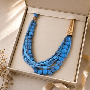 May include: A multi-strand necklace featuring various shades of blue beads in different sizes and textures. The strands are gathered at one end by a gold-toned metal cone with an etched pattern and at the other by a small gold-toned metal cap.