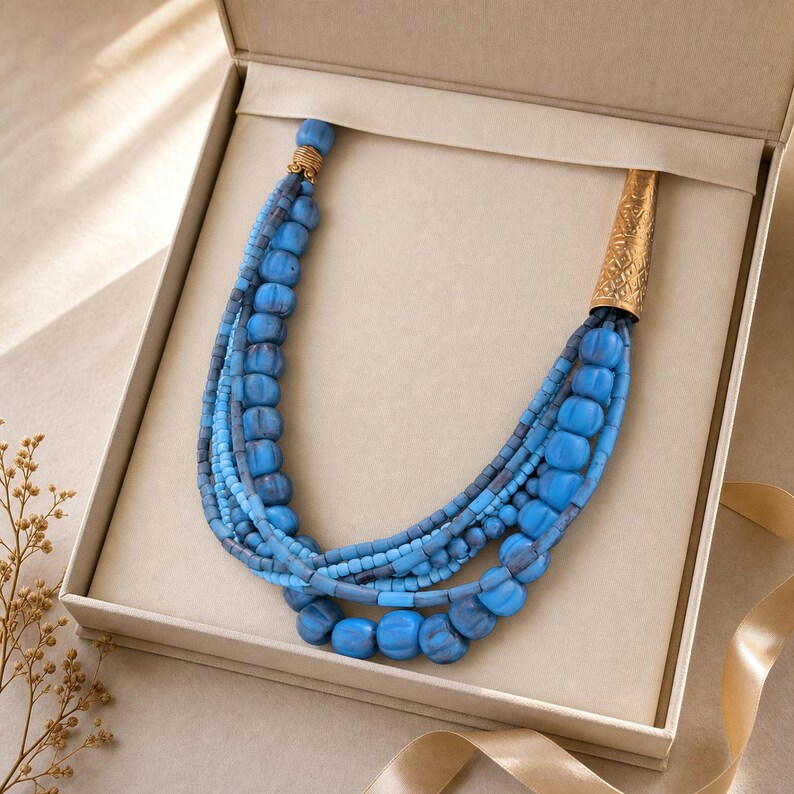 May include: A multi-strand necklace featuring various shades of blue beads in different sizes and textures. The strands are gathered at one end by a gold-toned metal cone with an etched pattern and at the other by a small gold-toned metal cap.