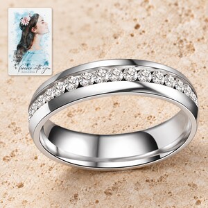 Close view of a silver-tone ring with one row of clear round clear accents and polished edges on pale travertine. A personalized digital greeting card is shown alongside.