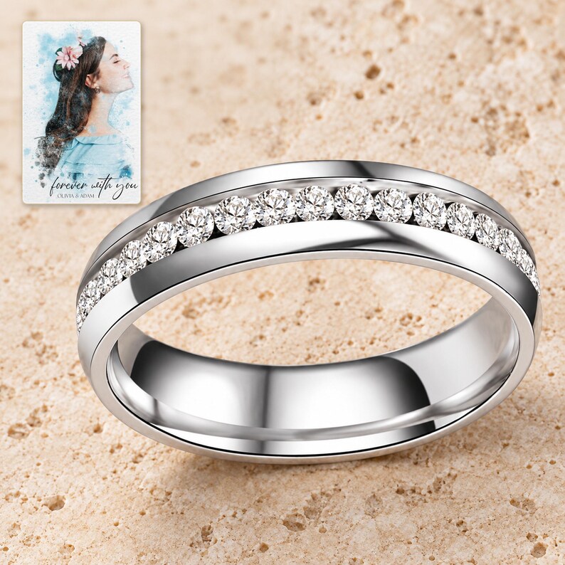 Close view of a silver-tone ring with one row of clear round clear accents and polished edges on pale travertine. A personalized digital greeting card is shown alongside.