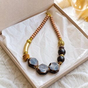 May include: A necklace featuring a strand of small light brown wooden beads, accented with gold-toned metal cylinders and spheres, dark brown round beads, and three large, polished, hexagonal dark brown marbled resin focal beads.