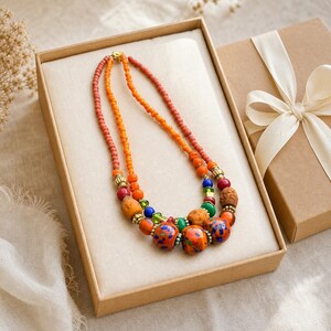 Peut inclure: Un collier &agrave; plusieurs rangs compos&eacute; de petites perles de rocaille corail et orange, agr&eacute;ment&eacute; de perles plus grosses orange, bleues, vertes et rouges, s&eacute;par&eacute;es par des intercalaires dor&eacute;s, pr&eacute;sent&eacute; dans une bo&icirc;te cadeau en carton beige.