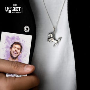 Puede incluir: Colgante de caballo plateado en una cadena fina. El colgante representa un caballo encabritado. La imagen tambi&eacute;n muestra un peque&ntilde;o retrato con el texto "GIFT ART DREAM DESIGN".
