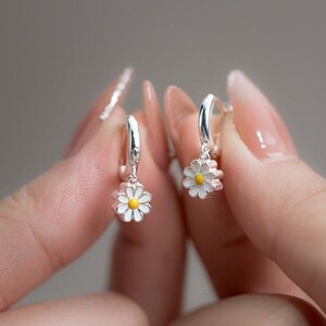 Pair of silver-tone hoop earrings with white daisy flowers and yellow centers held between fingertips.