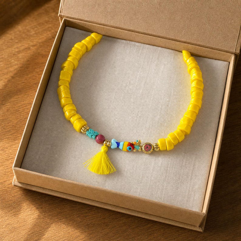 May include: A beaded necklace featuring bright yellow cylindrical beads, a small blue and orange fish-shaped charm, a red bead, a gold-toned circular accent, and a vibrant yellow tassel, all arranged in a semi-circular display inside a box.
