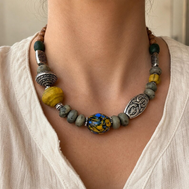Puede incluir: Un collar de cuentas con una cuenta central de vidrio moteado en azul y amarillo, flanqueada por cuentas de piedra gris, espaciadores de metal plateado texturizado y una cuenta amarilla grande. Incluye cuentas marrones y verde oscuro cerca del cierre.