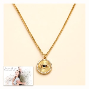 Gold-tone necklace with a round sunburst and black eye detail, catalog view. A personalized greeting-card example appears in the corner.