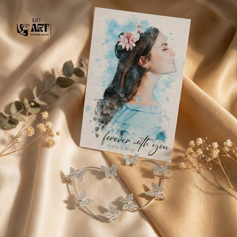 May include: A silver butterfly ear cuff with clear crystals and a greeting card featuring a watercolor portrait of a woman. The card reads "forever with you" and "Olivia & Adam". The image is set against a satin background.