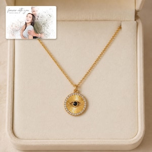 Gold-tone necklace with a round sunburst and black eye detail, presentation in an open jewelry box. A personalized greeting-card example appears in the corner.