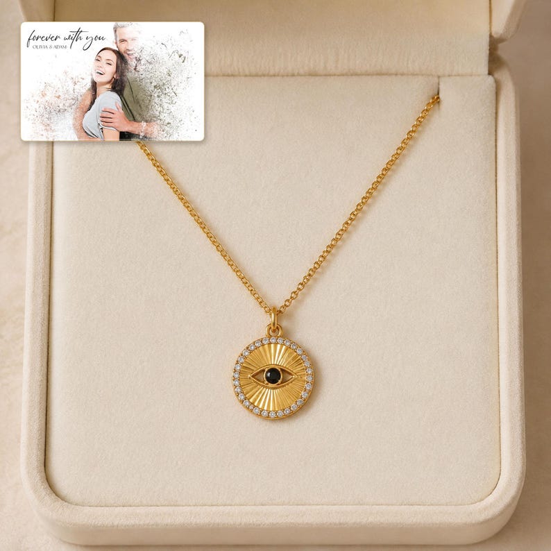 Gold-tone necklace with a round sunburst and black eye detail, presentation in an open jewelry box. A personalized greeting-card example appears in the corner.