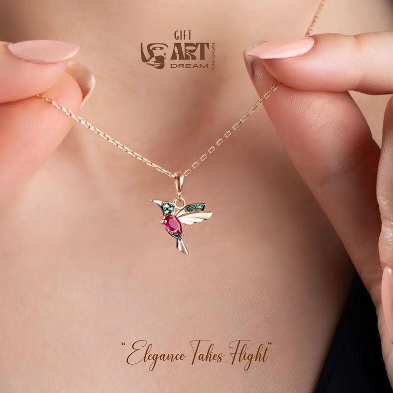 May include: A gold-colored necklace with a hummingbird pendant. The hummingbird pendant features a red gemstone body, green gemstone accents, and white wings. The necklace is held by a person. The text "Elegance Takes Flight" is at the bottom.