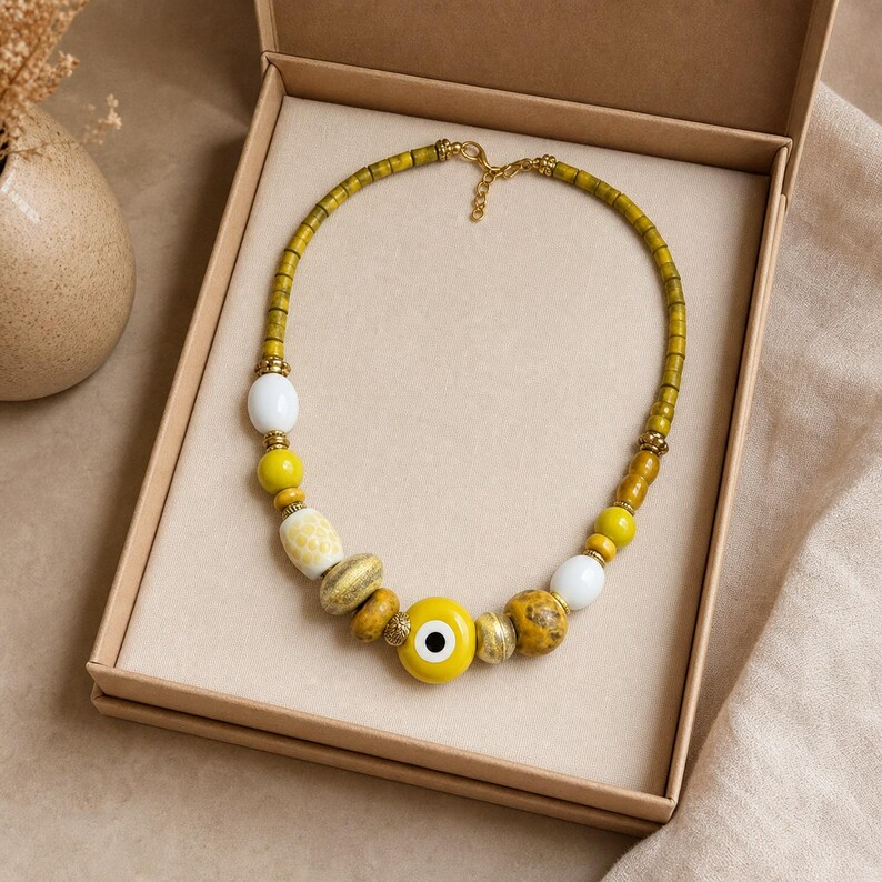 May include: A beaded necklace featuring a central yellow evil eye charm, interspersed with various yellow, white, and mottled brown beads. The necklace includes small gold-toned metal spacers and a gold-toned clasp closure, presented in a box.