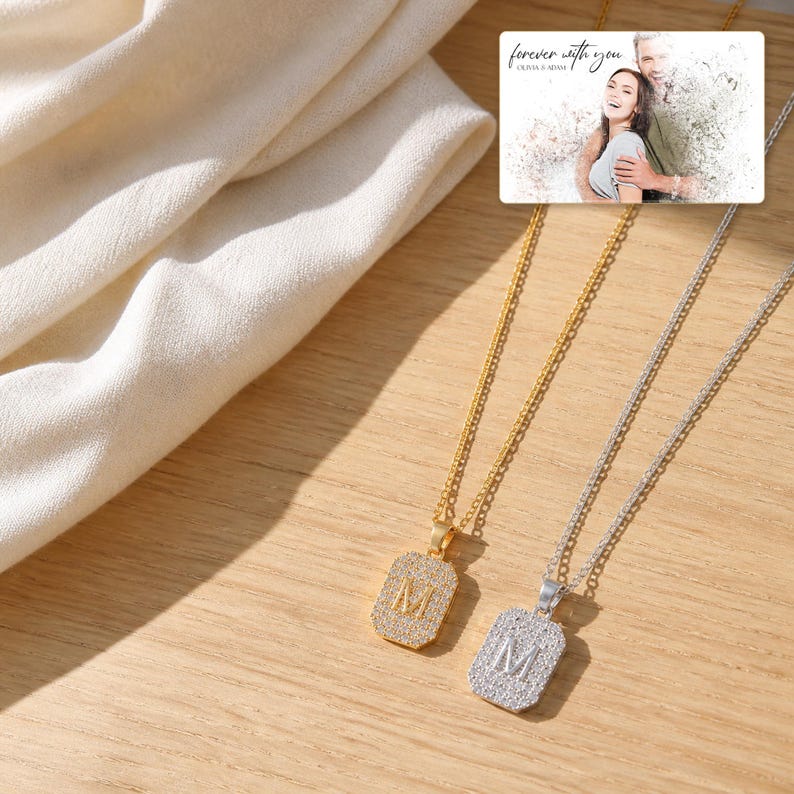 May include: Two rectangular pendant necklaces, one gold-toned and one silver-toned, each featuring a central letter M encrusted with small clear stones. A small rectangular card is visible with the text "forever with you" and "OLIVIA & ADAM".