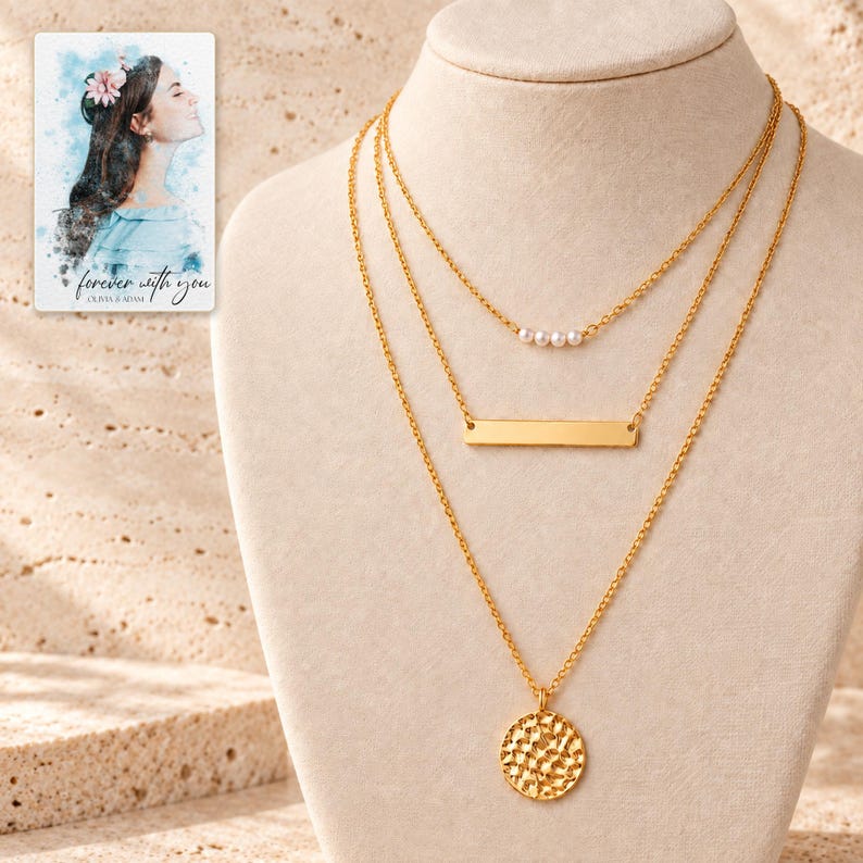 Three gold-tone necklaces displayed large on ivory fabric: four small white simulated pearl-style beads, a wide smooth bar and a hammered round coin. A personalized digital greeting card is shown alongside.