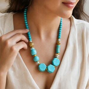 May include: A necklace featuring turquoise-colored beads in various shapes, including round, cylindrical, and hexagonal forms. The design incorporates ornate gold-toned metal spacers between the beads, creating a structured arrangement around the neck.