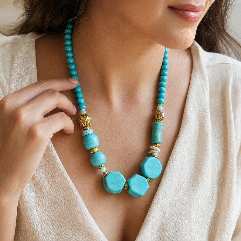 May include: A necklace featuring turquoise-colored beads in various shapes, including round, cylindrical, and hexagonal forms. The design incorporates ornate gold-toned metal spacers between the beads, creating a structured arrangement around the neck.